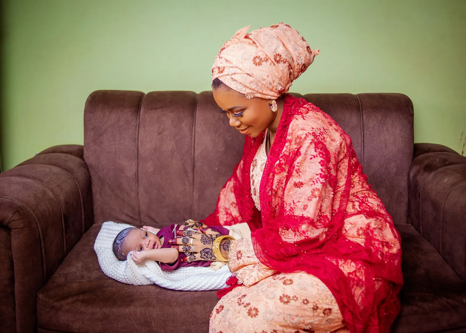 Nigerian mother in a coral lace outfit and headwrap smiling down at her newborn on a sofa