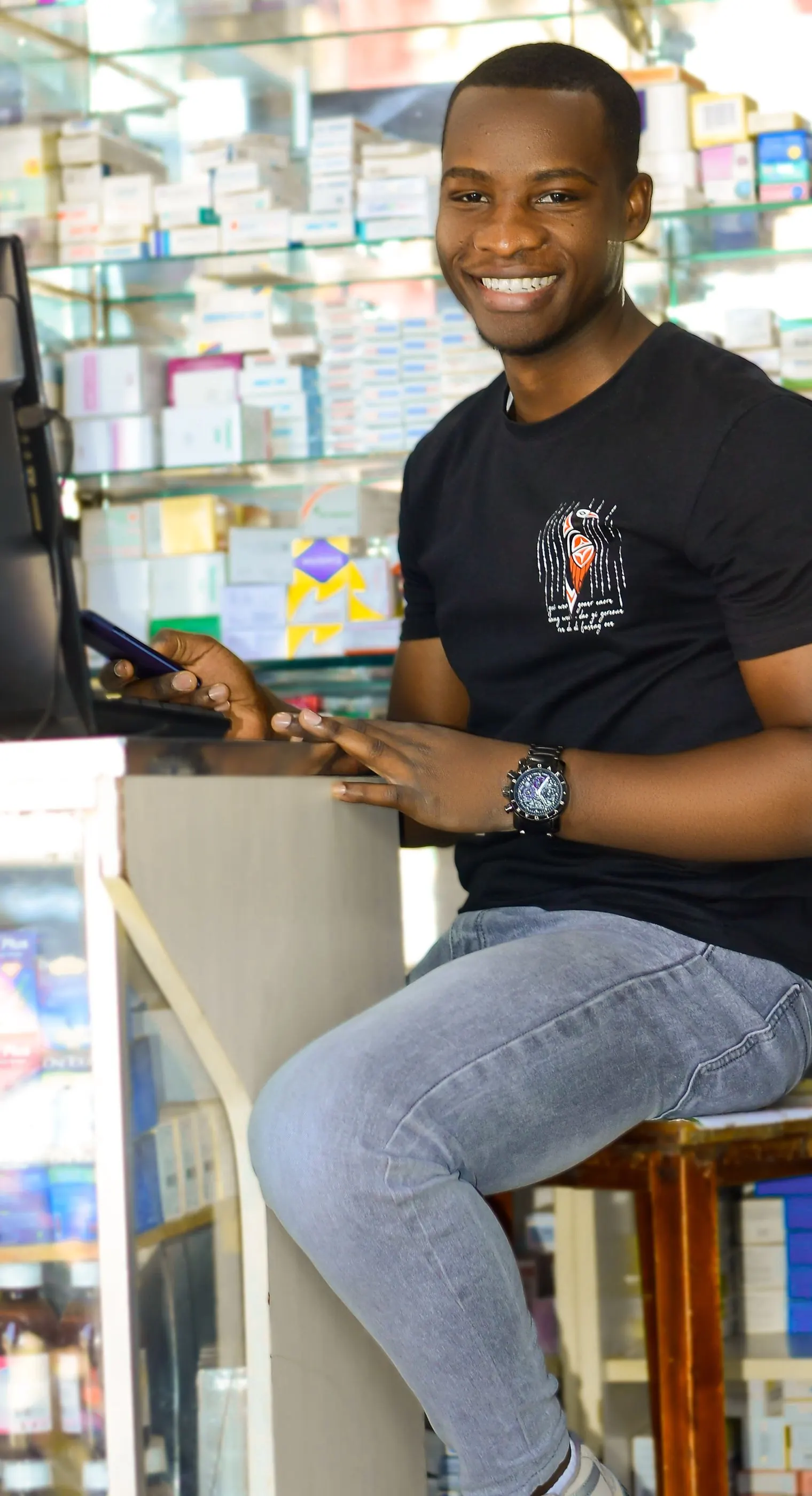 Smiling pharmacy attendant seated at the counter in front of shelves of medicines